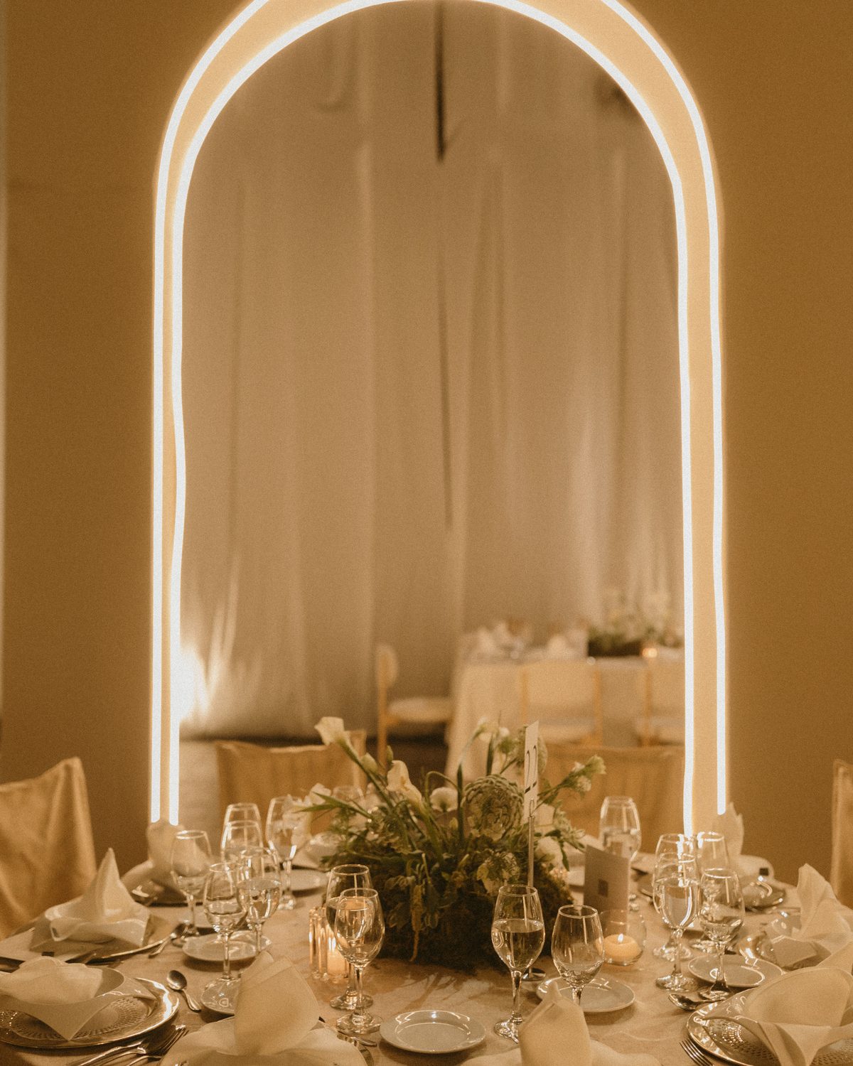 Una mesa redonda con flores blancas y velas, enmarcada por un arco iluminado y cortinas claras.