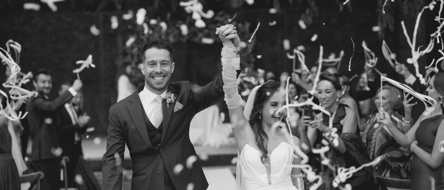 Una pareja sale bajo una lluvia de confeti blanco mientras sus invitados celebran a su alrededor.