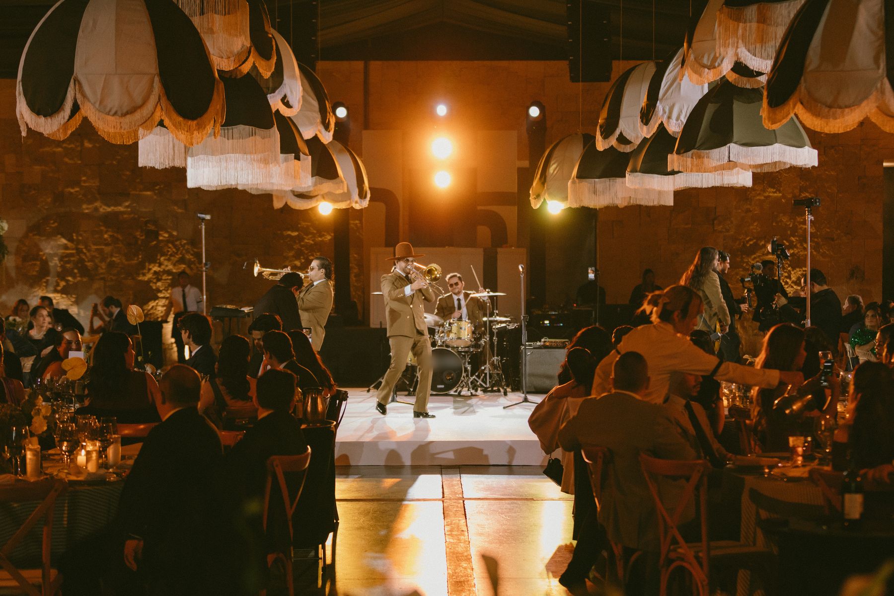 El salón durante la cena, visto desde atrás de los invitados: todas las mesas llenas y la banda tocando en un escenario iluminado bajo las lámparas de flecos.