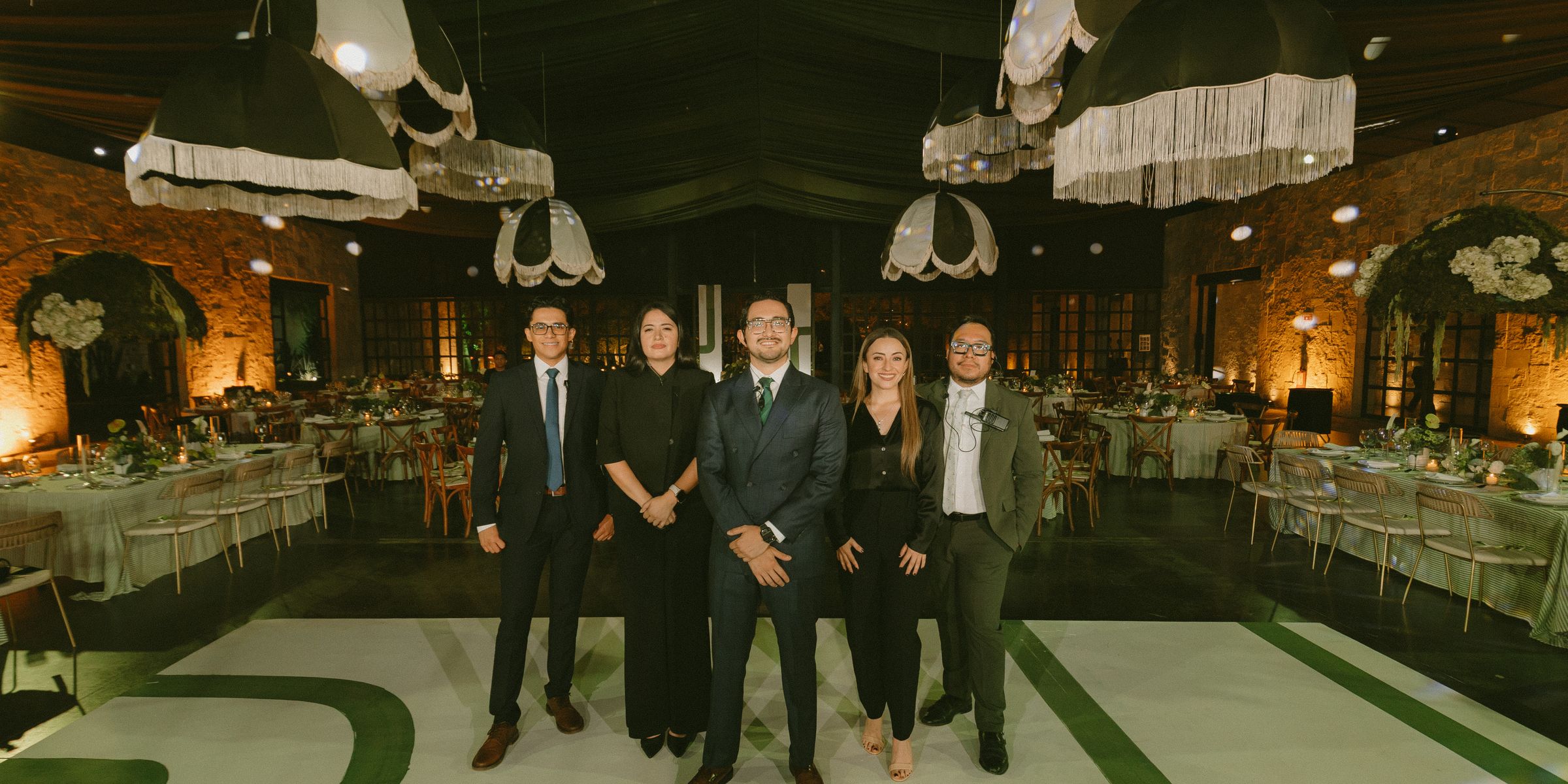 El equipo de CMeraz Studio, cinco personas de traje oscuro con intercomunicadores, de pie sobre la pista de laberinto verde bajo las lámparas de flecos, con las mesas montadas detrás.