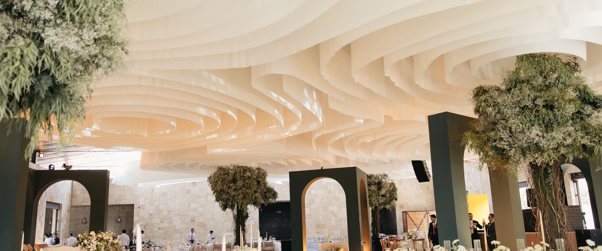 Vista hacia un techo blanco en ondas, con arcos olivo oscuro y árboles frondosos dentro del salón.