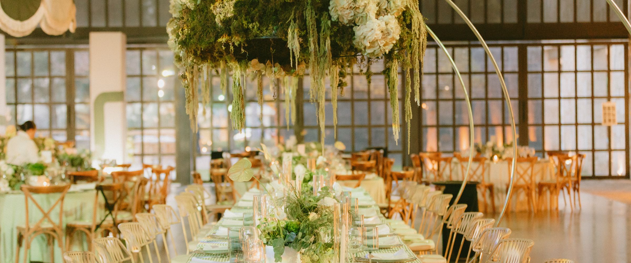 Una corona suspendida de hortensias blancas y follaje sobre una mesa larga vestida en salvia, con aros dorados y ventanales altos al fondo.