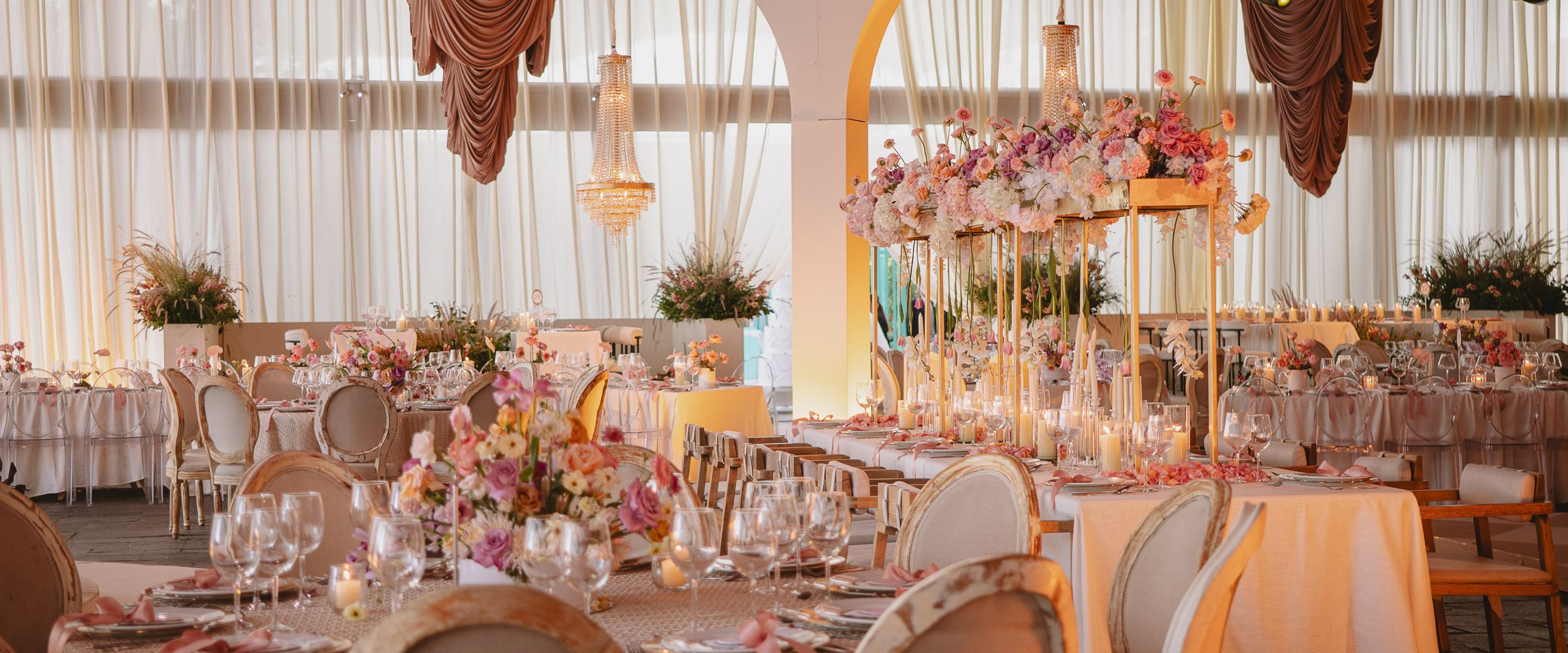 Un salón de recepción con luz suave de día: cortinas claras, un arco blanco, candiles de cristal y drapeados rosados sobre mesas llenas de flores rosas y coral.