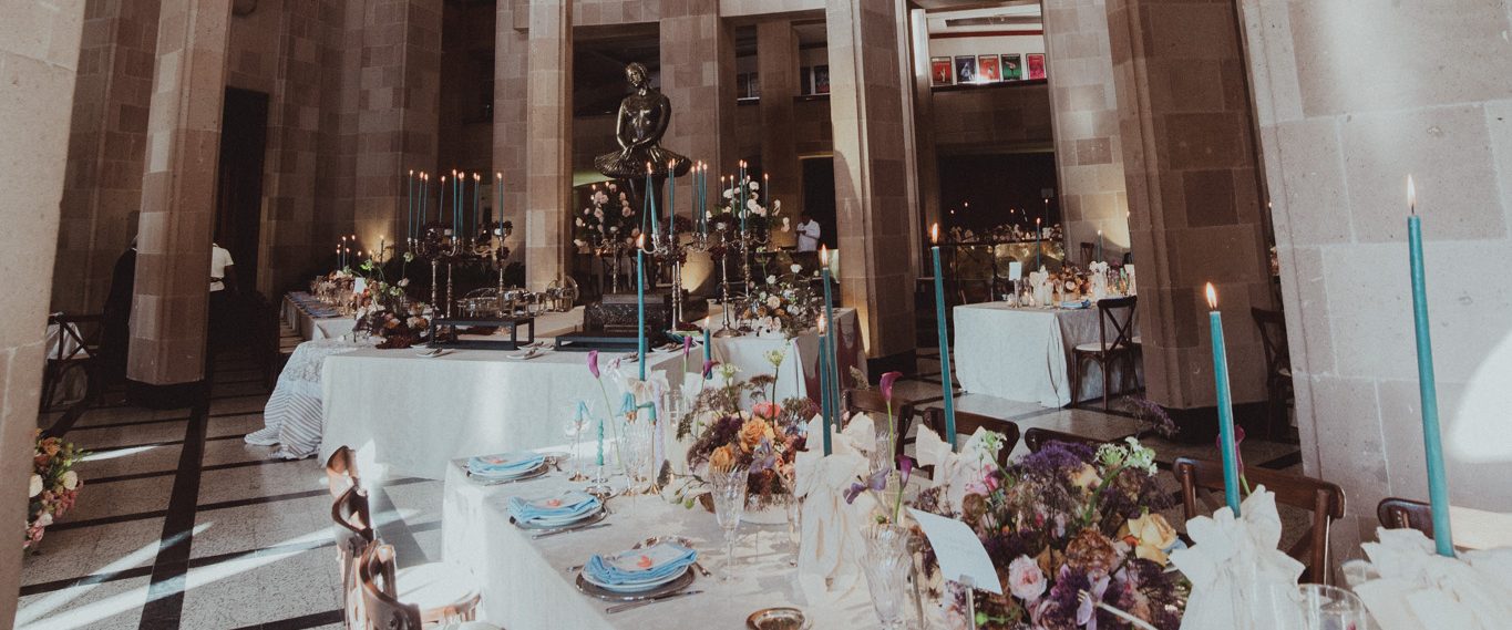 Un salón de cantera con cúpula, columnas altas y una escultura de bronce; una mesa larga vestida de blanco con velas altas color turquesa y servilletas azules.