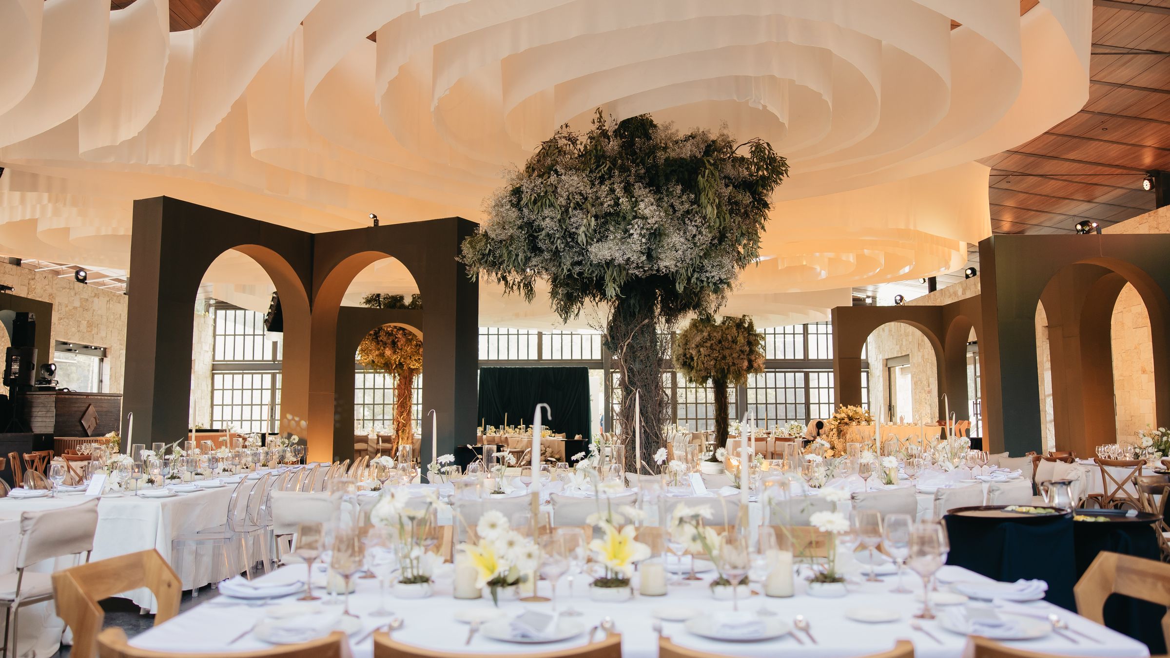 Un salón bajo un techo de tela blanca en ondas, con arcos olivo oscuro, un árbol suspendido y mesas largas vestidas de blanco.